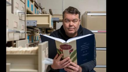 Researcher Mark Gwynn holds open a copy of the Australian National Dictionary, surrounded by filing cabinets.