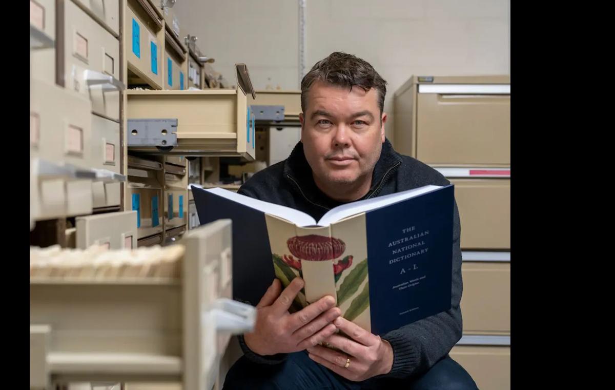 Researcher Mark Gwynn holds open a copy of the Australian National Dictionary, surrounded by filing cabinets.